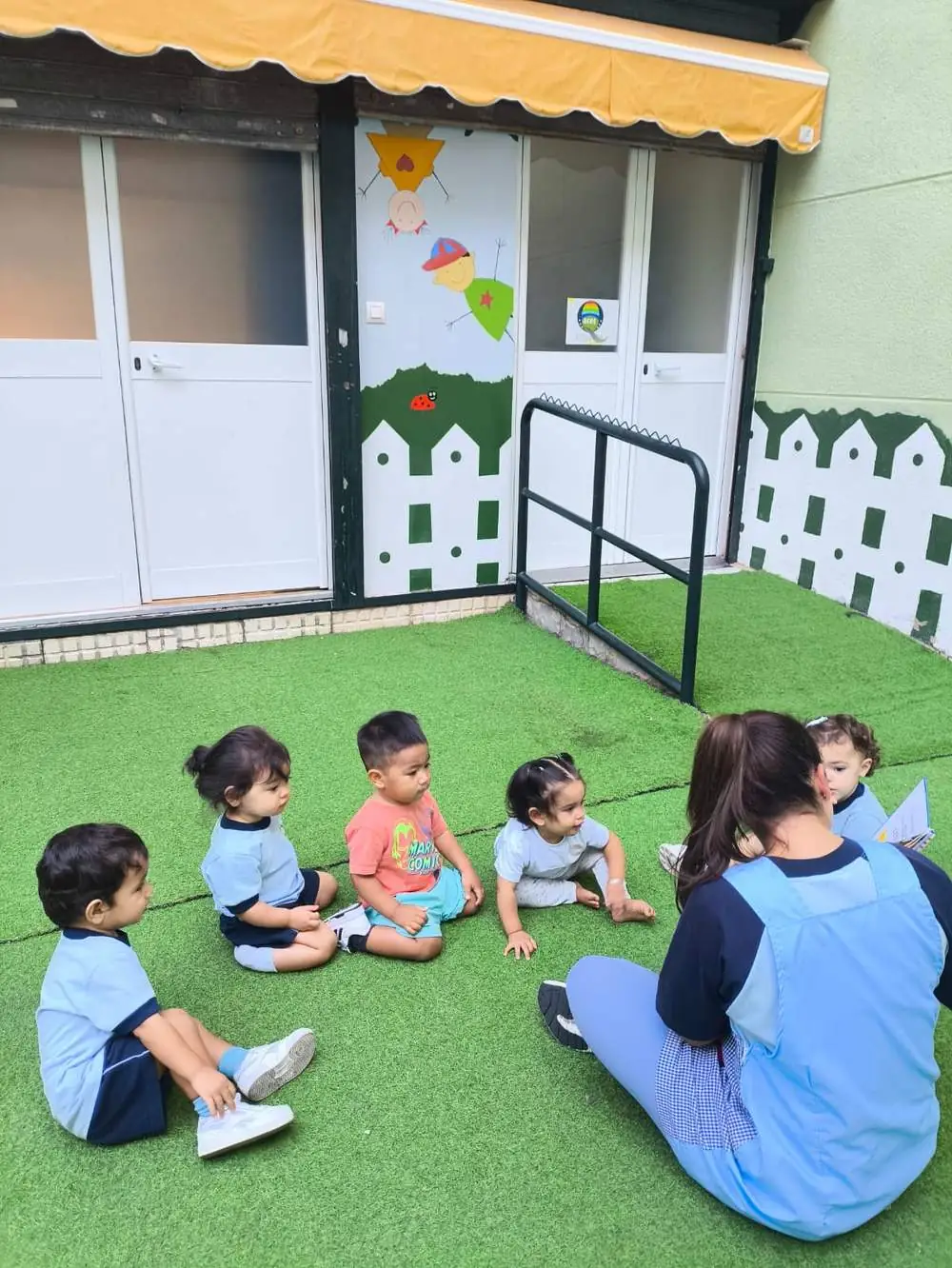 Peques sentados en el césped artificial del patio con su educadora
